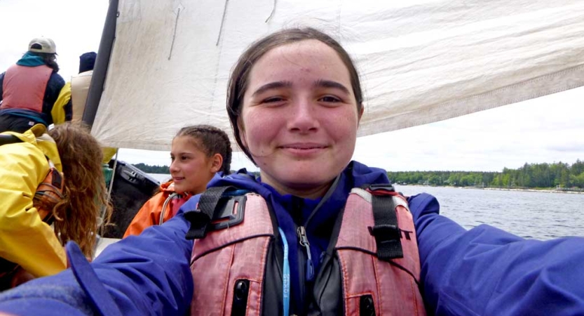 A student wearing a life jacket takes a selfie and smiles in front of a white sail